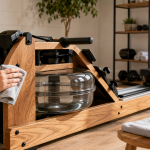 Hands wiping down the wooden frame of a WaterRower rowing machine with a microfiber cloth, with wood care oil nearby, in a bright home gym setting.